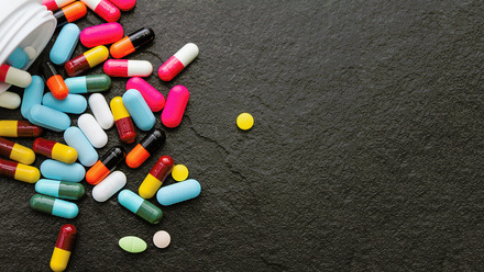 Image features a collection of multicoloured pills spilling out from container, on black counter.